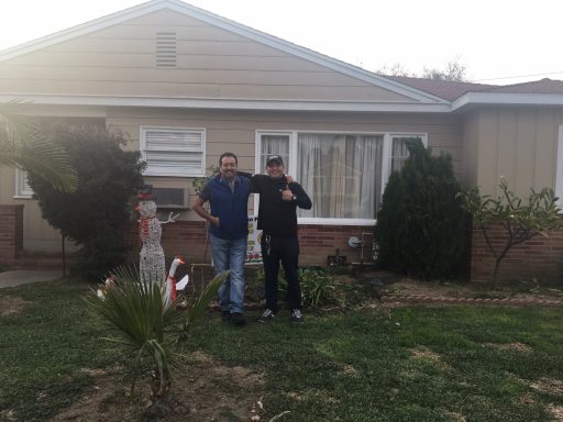 Two men stand outside a house, posing near a small palm tree and garden area.