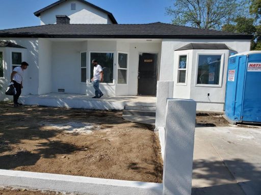 Newly constructed house with a front porch, two workers, and a portable restroom nearby.